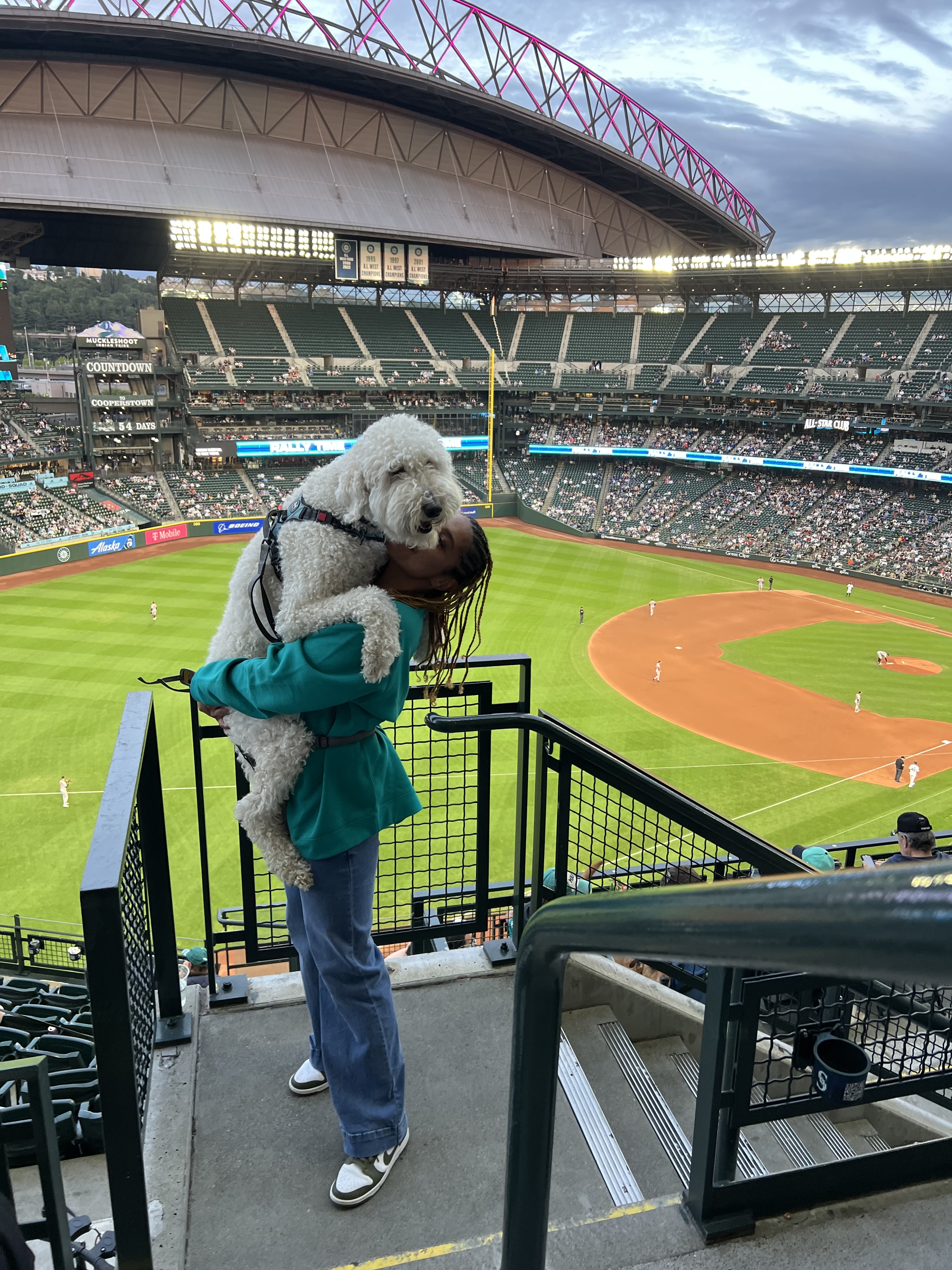 Brianna stands in a baseball stadium holding her large white fluffy dog in her arms while smiling; the field and crowd are visible below.