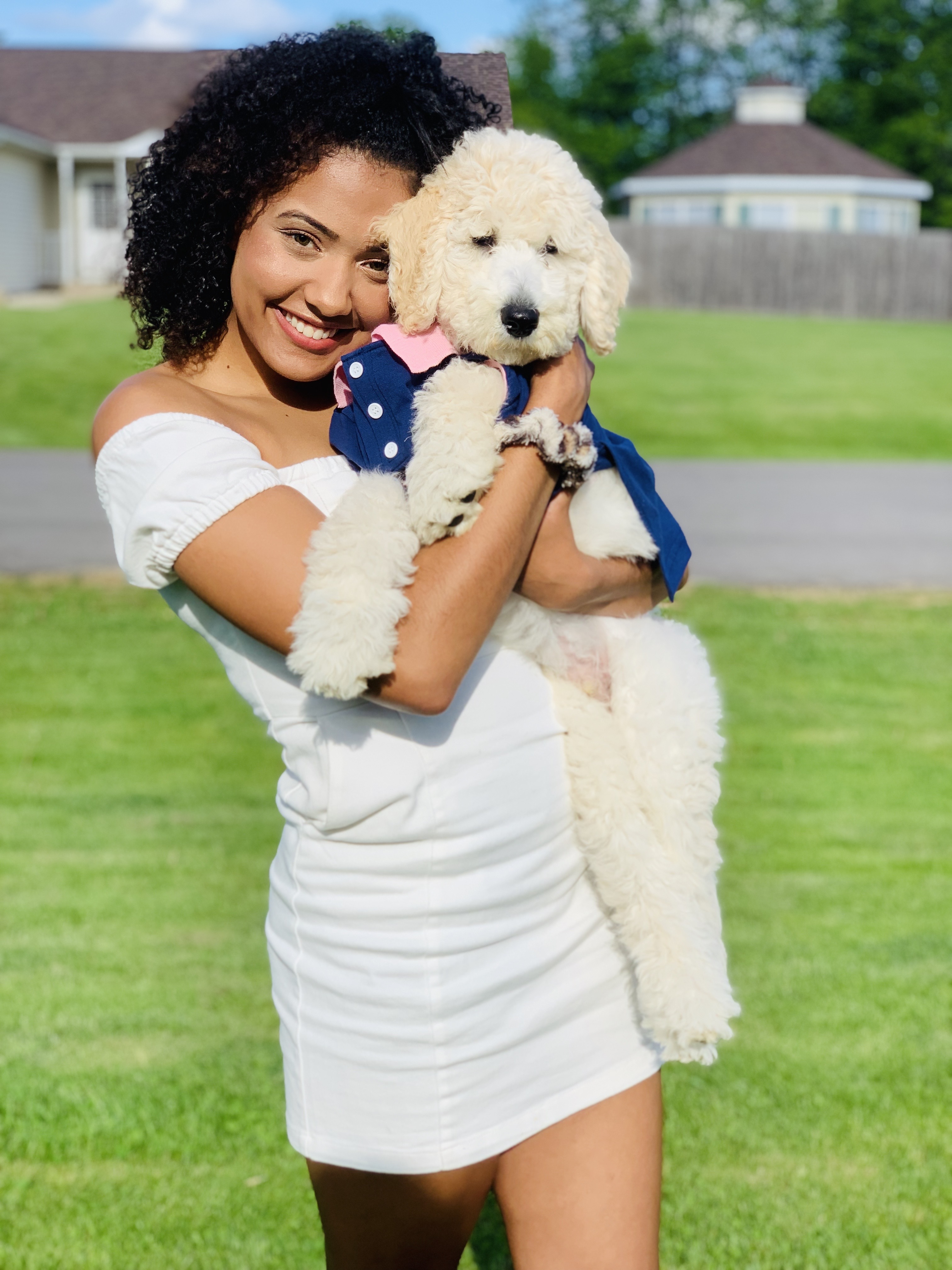 African American woman in a white dress holding a white puppy, which is wearing a blue and pink shirt.