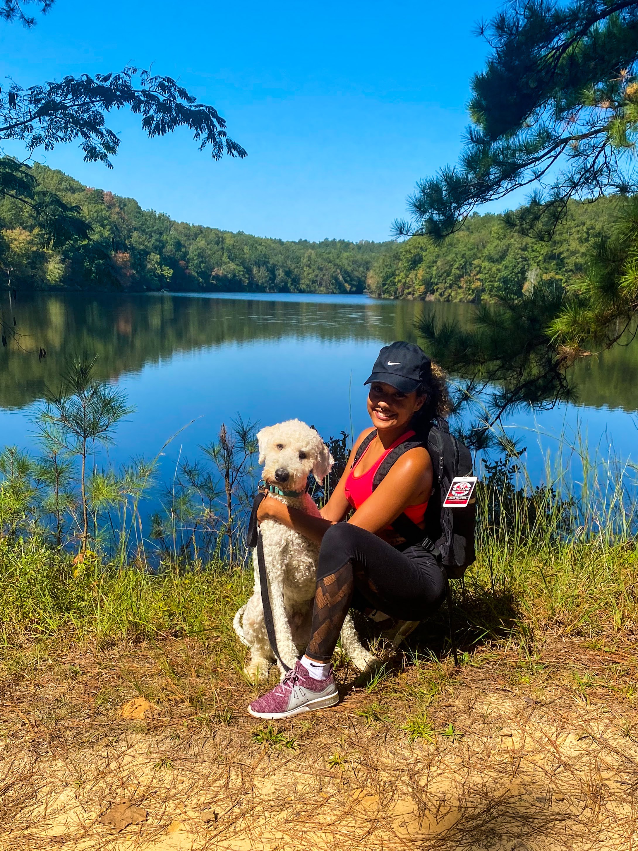 African American woman squatting beside a white dog, embracing it, with a scenic lake in the background.