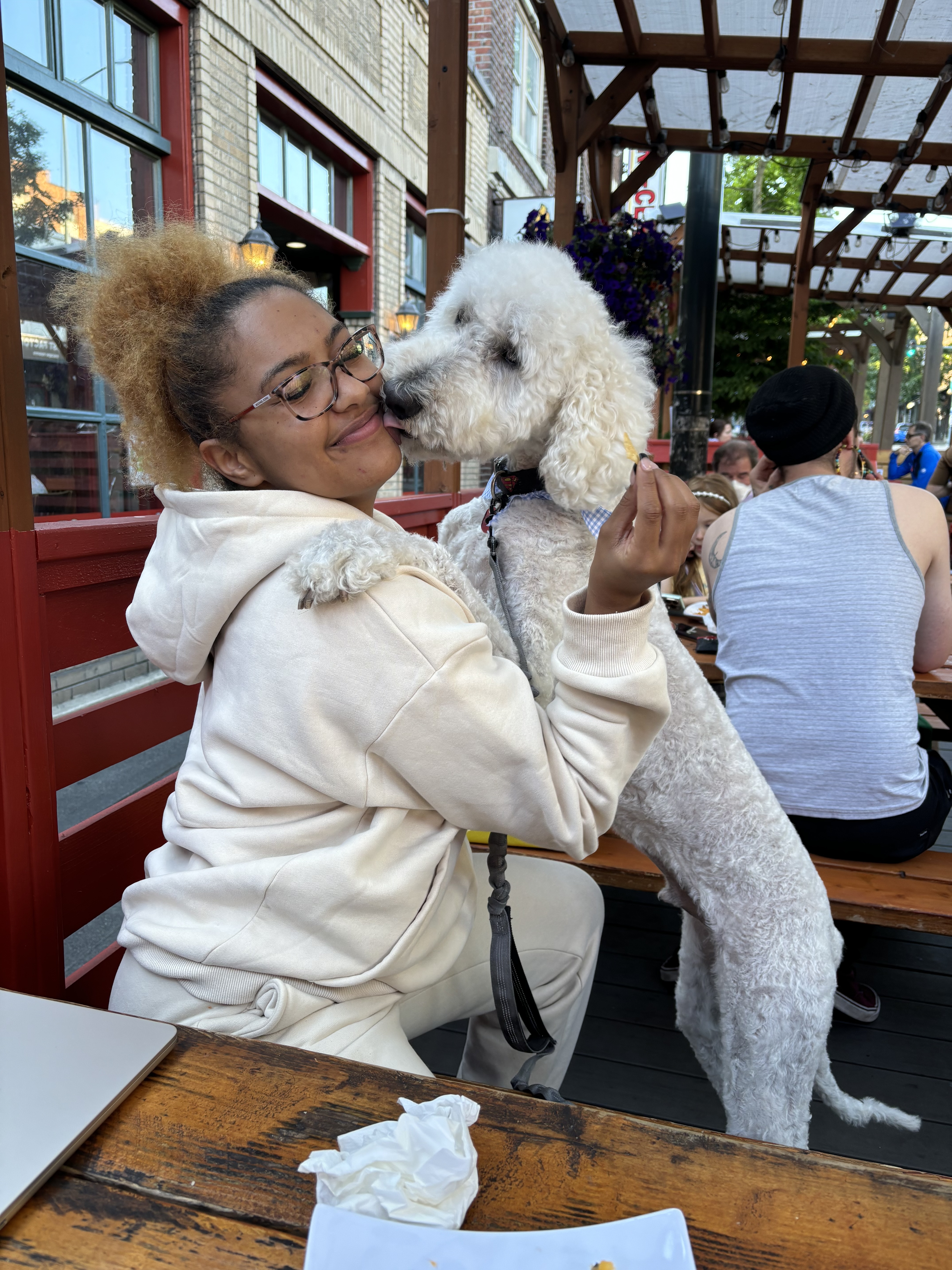 Brianna sits at an outdoor restaurant table as her white dog leans in to lick her face; she’s laughing and wearing a cream-colored sweatsuit.