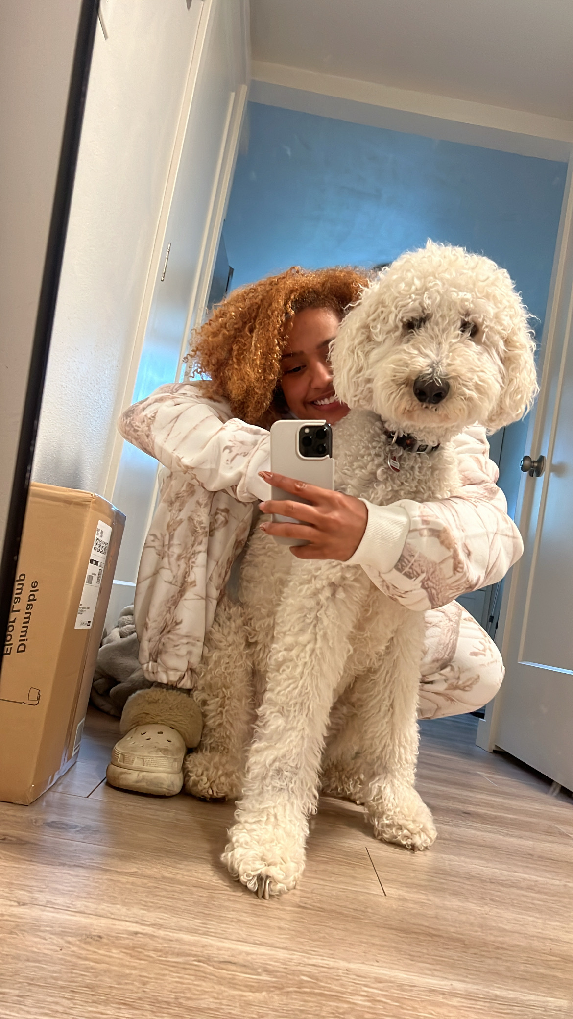 Brianna kneels on the floor hugging her white fluffy dog in front of a mirror, smiling while taking a selfie together.